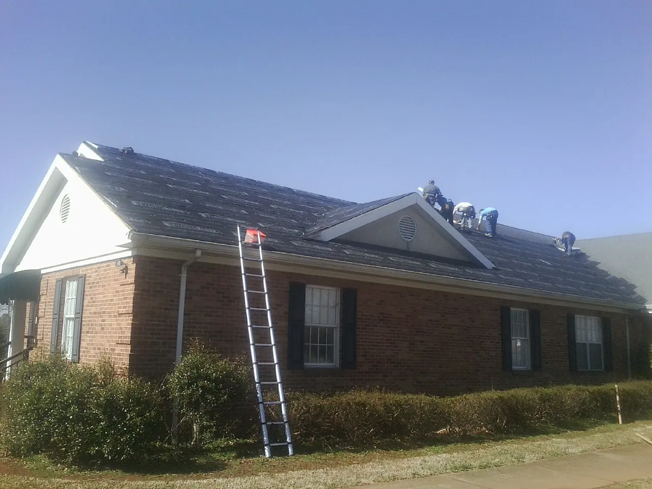 Crew tearing shingles off a brick ranch roof with a ladder set — Emergency Roof Repair work by our crew in Pittsboro
