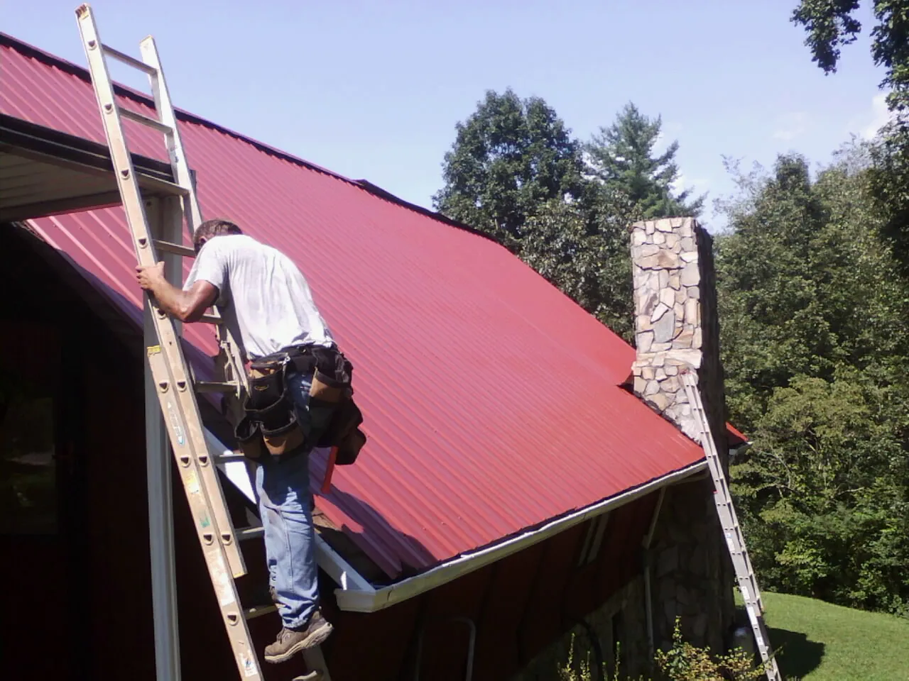 Roofer on a ladder beside a red metal roof and stone chimney — Soffit & Fascia Repair by Roofing Brothers in Pittsboro